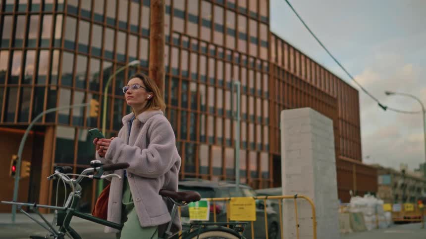 A woman in a light gray fleece coat on a bicycle on a city street is looking at her smartphone with earbuds in