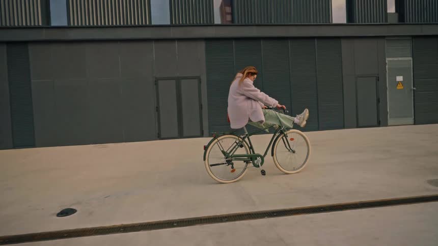 A woman in a light gray coat rides a green bicycle playfully with both legs stretched straight out in front of her gliding across a wide empty concrete space in front of a modern industrialstyle building