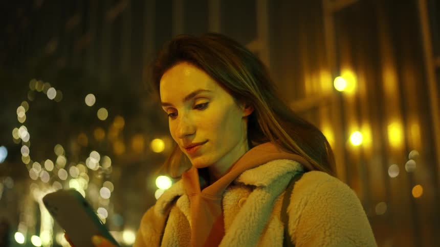 A woman focuses on her smartphone amid the warm glow of nighttime city streets