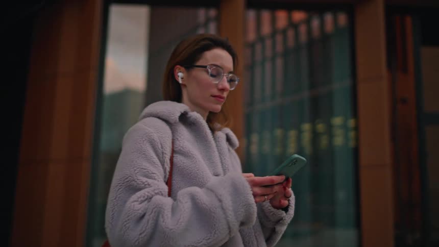 A woman wearing glasses and white earbuds walks past a modern glass building while holding a smartphone she is wearing a light grey fuzzy coat and carrying a large reddishbrown bag over her shoulder