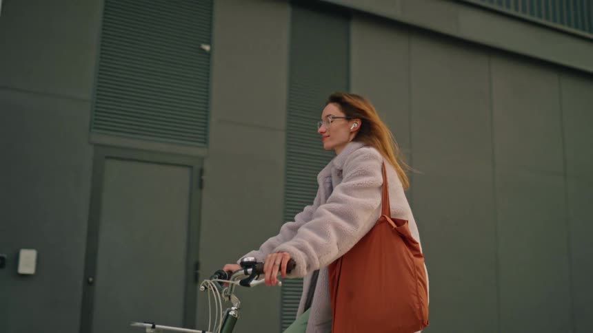 Woman rides her bicycle on the sidewalk in front of buildings moving her head to the music from her earbuds