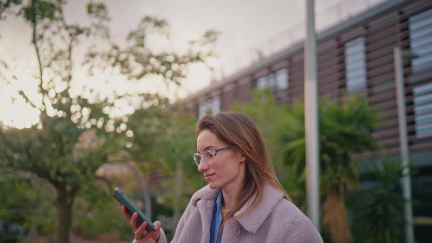 Outdoors near a modern slatted building a woman in a plush lightgray coat and glasses pauses with a bicycle glancing at a teal smartphone amid trees palmlike plants and a lamppost at dusk