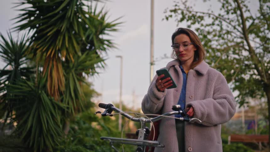 Woman holding a bicycle while using a smartphone in a treelined outdoor area