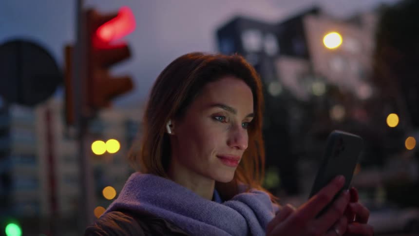As the camera orbits a woman standing on a city street corner at dusk the soft glow from her phone illuminates her face against the blurred backdrop of evening traffic lights