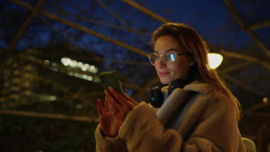 A woman wearing glasses and a fuzzy jacket stands outdoors at night while looking at her smartphone warm streetlights glow in the background as she smiles gently and scrolls through the content on her screen