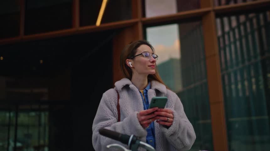 A woman with glasses and a purple fleece coat stands with her bicycle in front of a modern building while using her mobile phone and looking around at dusk