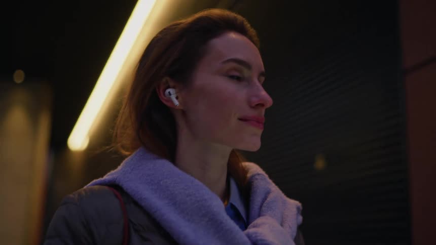 Woman using a smartphone with wireless earbuds in a nighttime urban setting
