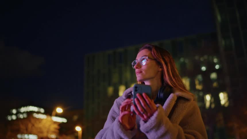 A young woman checks her phone while standing on city streets outdoors at night