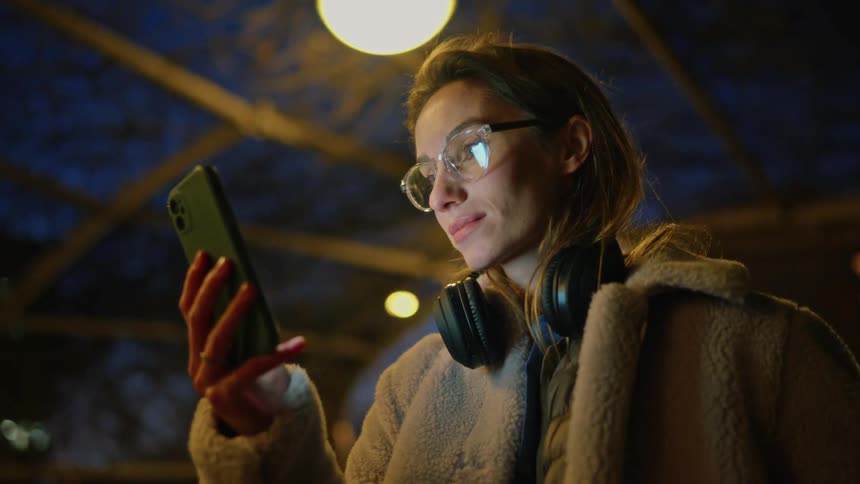 A woman with glasses and a lilac fleece coat stands on a city street at night while holding her phone and wearing headphones around her neck under warm streetlights