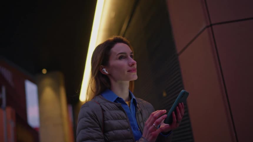 Woman using a smartphone with wireless earbuds in a nighttime urban environment