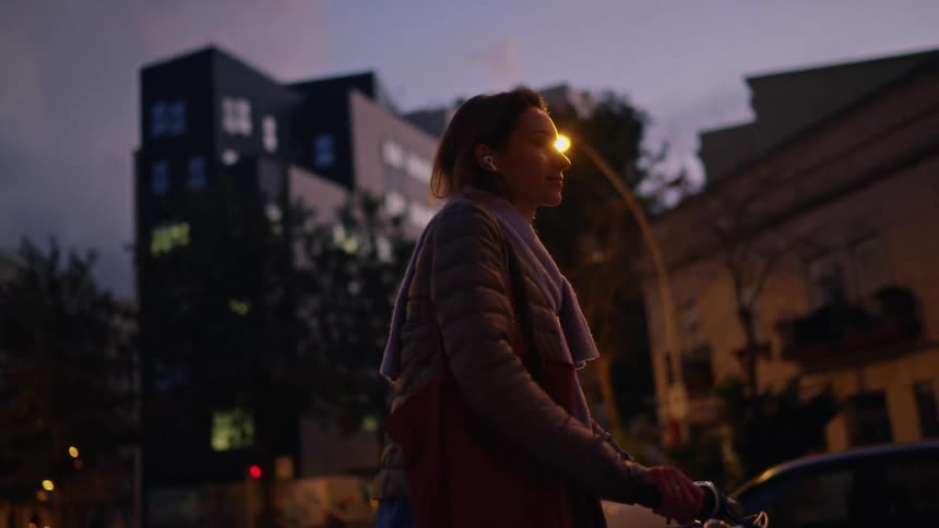 A woman on an bike rides down a quiet city street at dusk the camera slowly closing in on her thoughtful expression as she passes under warm streetlights
