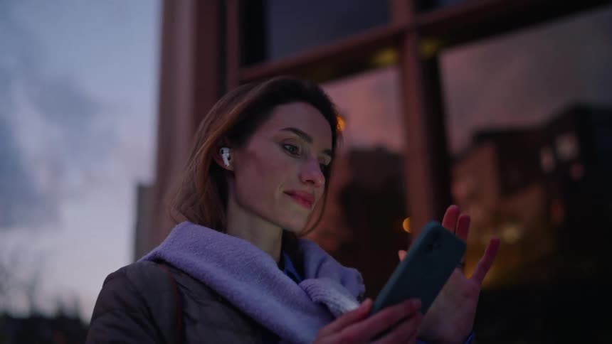 Woman using a smartphone with wireless earbuds in a nighttime urban environment