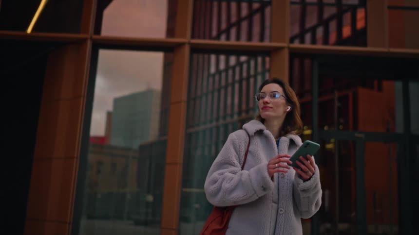 A woman checks her smartphone while walking outside a modern city building