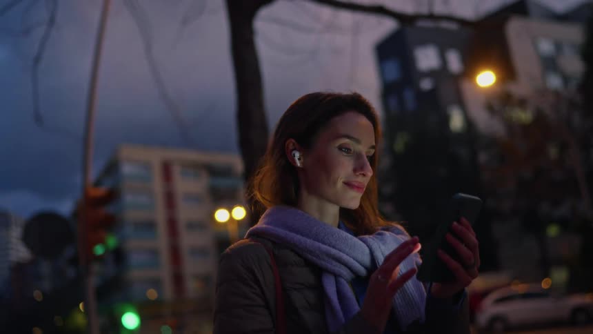 A woman wearing a brown jacket and a light blue scarf stands on a city street at night while looking at her phone with wireless earbuds in her ears