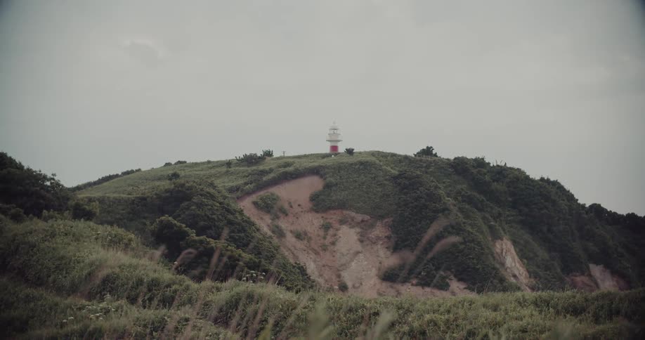 A distant red and white lighthouse sits atop a vast green hill under an overcast sky while tall grass in the foreground gently sways in the wind