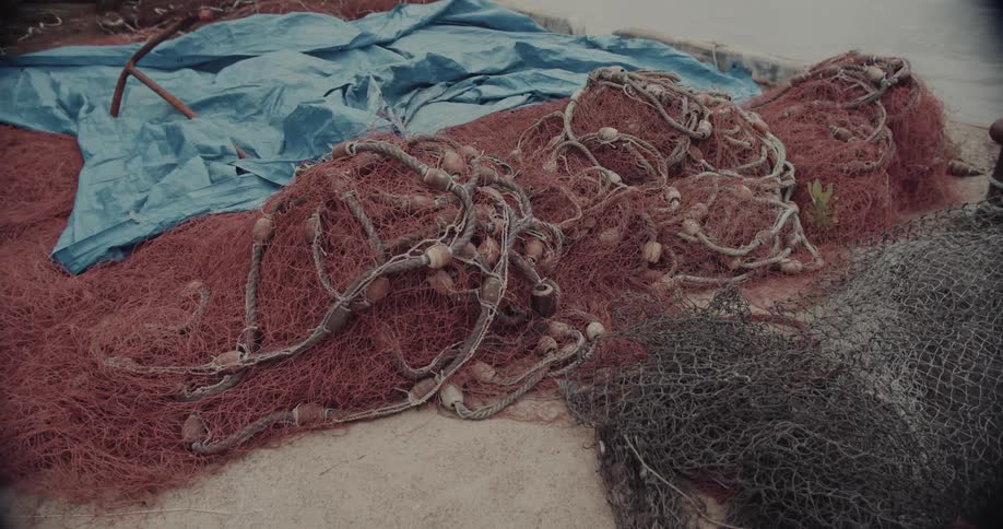 A pile of tangled reddishpink and black fishing nets rests on a flat weathered surface in the background a blue tarp and a rusty metal anchor sit near the edge of the water