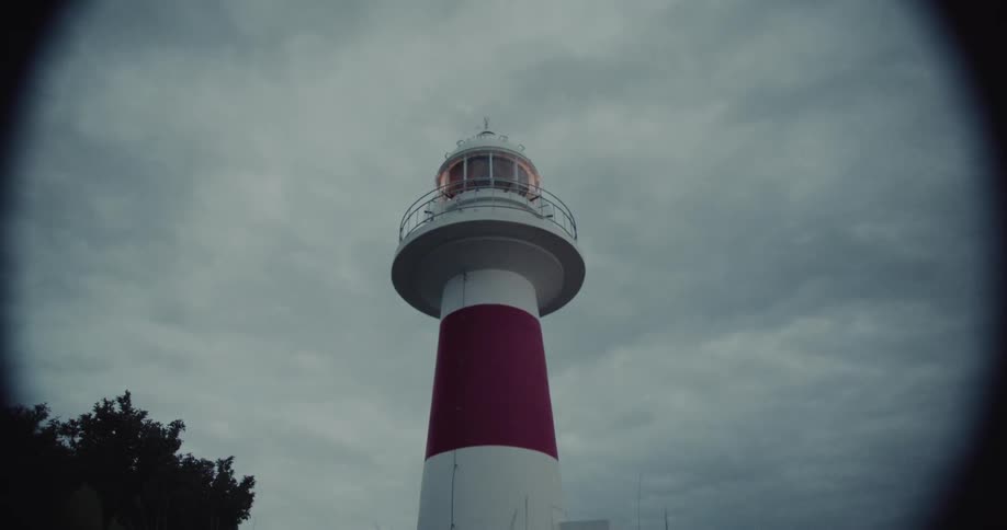 A tall red and white striped lighthouse stands out against a heavily clouded dark sky with its lamp illuminated