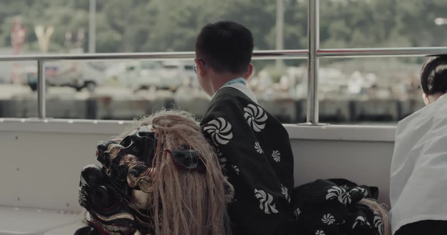 A child dressed in a patterned black traditional robe sits on a boat beside an ornate lion dance head and a woman gazing out through metal railings toward a busy harbor and treelined shore