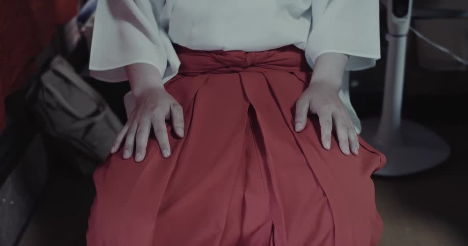 A woman sitting indoors wearing a white top and a red skirt both hands resting neatly on her lap