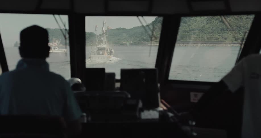 From the shadowy wheelhouse of a ship two sailors watch another boat as it moves across the water toward a distant treecovered coastline