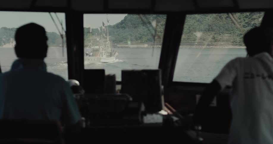 From the bridge of a vessel two crew members look ahead through large windows at another boat traveling in the same direction