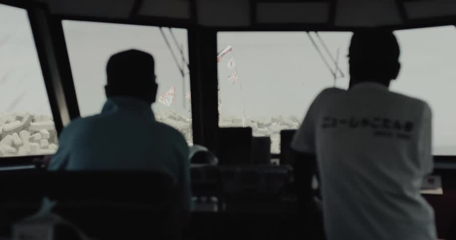 In the bridge of a vessel a pilot sits at the helm while another crew member stands nearby both looking out toward the wateroutside the large windows colorful flags flutter in the distance near a concrete breakwater and a red lighthouse