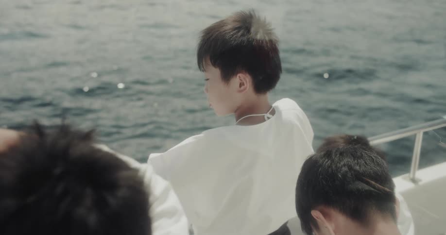 A child in a white shirt is standing on a boat looking out at the sparkling blue green water with the heads of other children partially visible around them