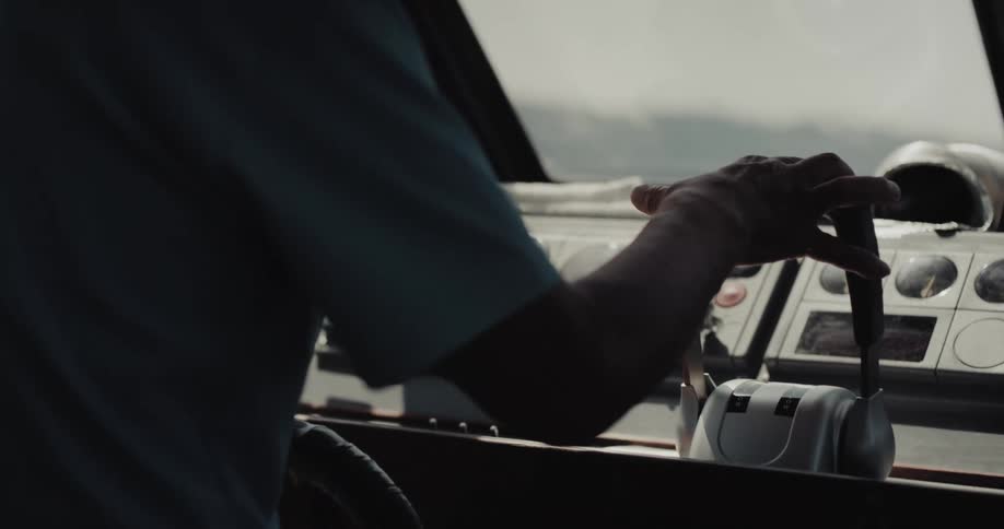 Inside the dim cabin of a vessel a captain rests his hand on the dual throttle levers adjusting the speed as the boat glides across the water under a bright midday sun