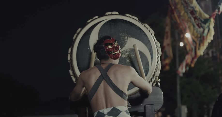 Masked person playing traditional japanese drum during nighttime festival with banners in the background
