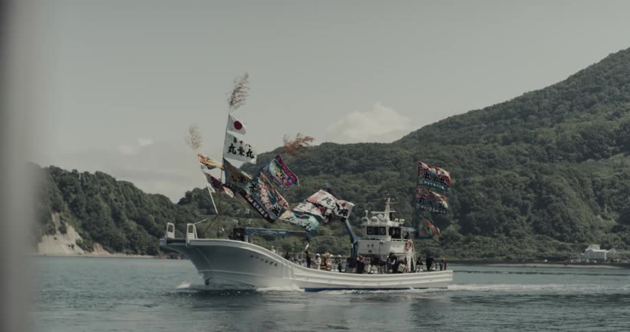 A white fishing boat decorated with colorful japanese flags travels steadily across the water set against a backdrop of steep forested mountains