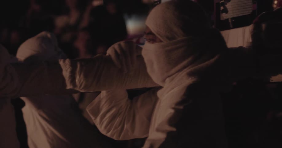 Two individuals in white clothing holding ceremonial object in dark setting with audience in background