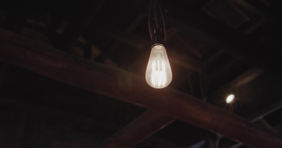 A single vintage filament bulb hangs suspended from a rustic wooden ceiling casting a steady warm amber glow into the surrounding darkness