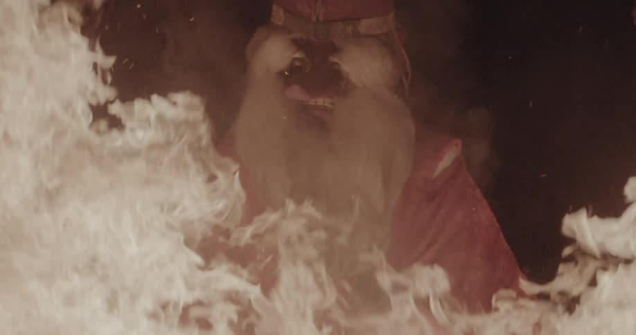 An elderly man with long grey hair wearing a mask dancing near a large fire yellow flames lighting up the night outdoors