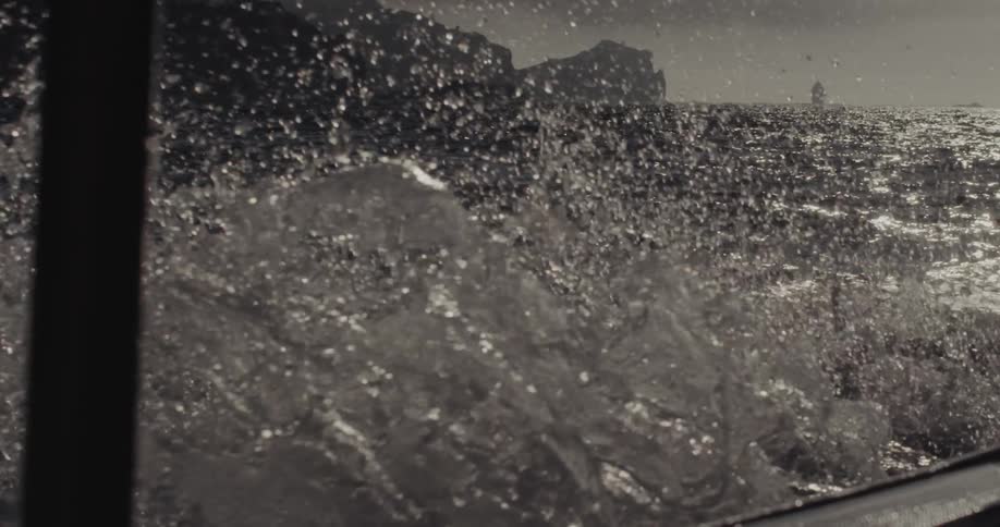 Sea water splashes forcefully against the side of a moving boat revealing a glimpse of distant rocky cliffs and sunbeams reflecting off the dark choppy waves as the vessel travels forward