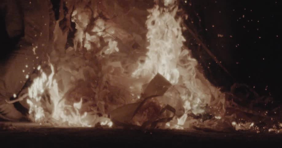 A pile of material burns intensely in the dark with a person standing nearby