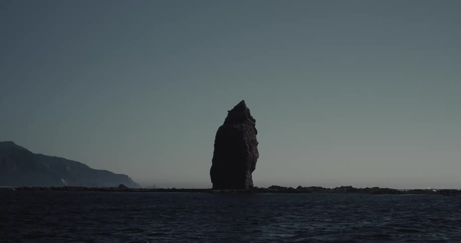 A massive jagged rock formation rises abruptly from the dark choppy waters standing as a stark silhouette against the moody sky while a small drone flies low near the surface