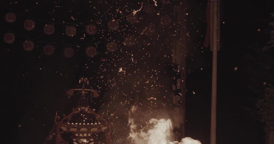 A large bonfire erupts with white flames and sparks at night near an ornate structure under a string of illuminated lanterns