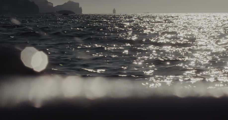 The camera captures a highcontrast view of shimmering ocean waves under a bright sun with the silhouettes of jagged mountains and a lone sea stack appearing on the distant horizon behind blurry foreground light flares