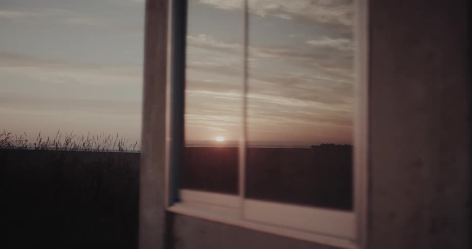 A window frames the setting sun reflecting on its glass situated above dark grasses with a muted hazy landscape beyond