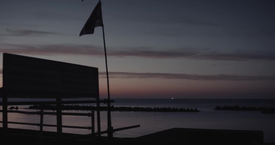 A dark flag flutters in the steady breeze atop a tall pole at dusk overlooking a quiet shoreline where a stone breakwater sits in the calm water under a fading purple and orange sky