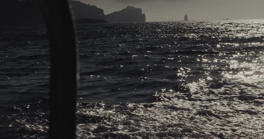 Sunlight glimmers across the churning ocean waves as a boat speeds past a rugged mountainous coastline with dramatic cliffs and a distant sea stack under a heavy overcast sky