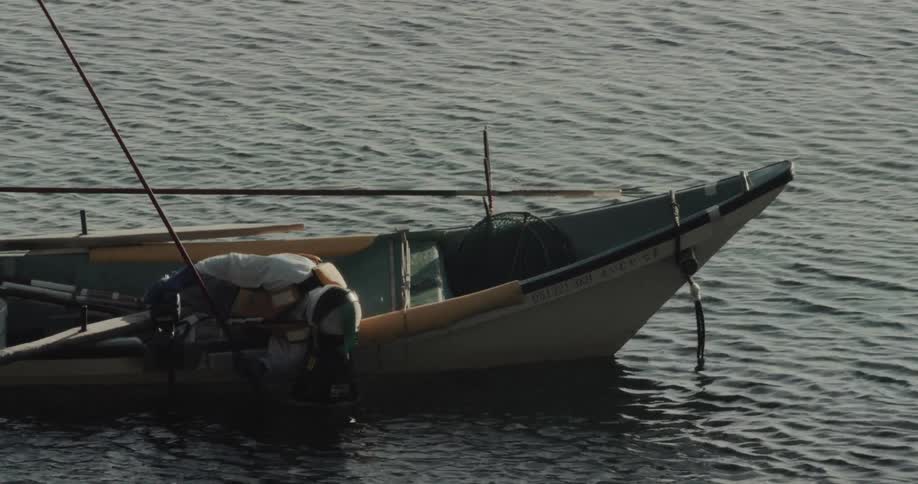 A fisherman wearing protective gear leans over the side of a small traditional fishing boat to inspect an outboard motor while the vessel floats on calm open water during the day