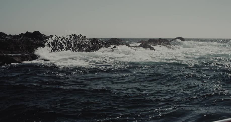 Waves crash powerfully against dark jagged rocks emerging from the deep blue choppy sea under a bright pale sky