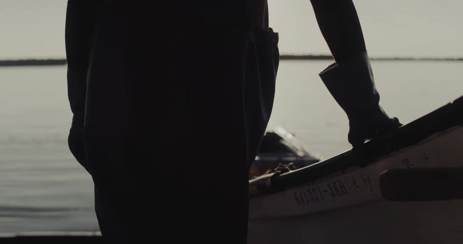 A figure in dark clothing leans against the side of a boat at the waters edge during bright hazy daylight
