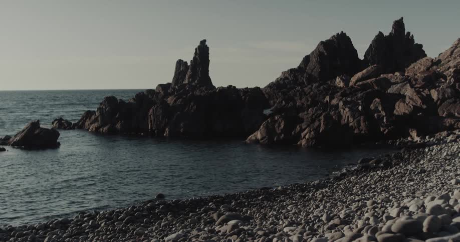 Large jagged rocks stand along a pebblecovered shore while dark ocean waves gently wash against the coastline under a overcast sky
