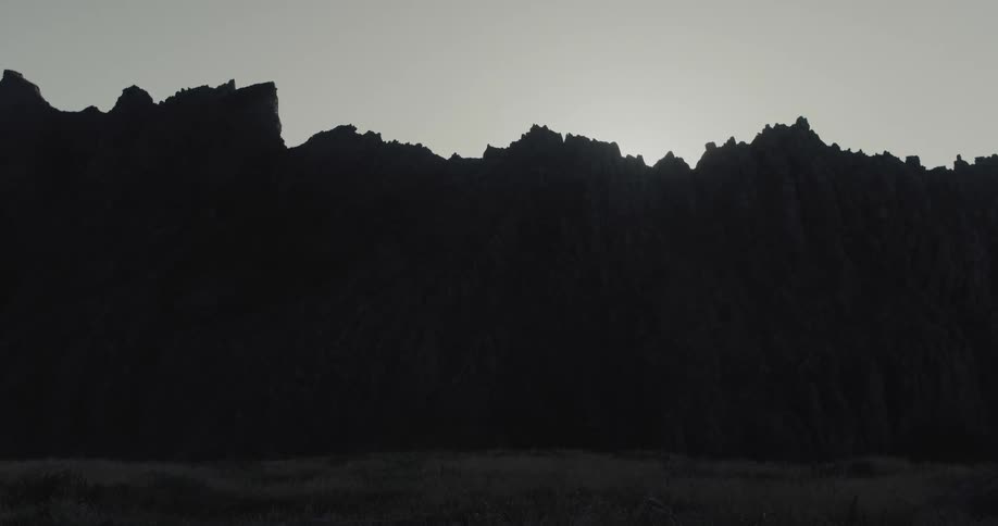A jagged mountain ridge is silhouetted darkly against a bright overcast sky above a muted foreground landscape