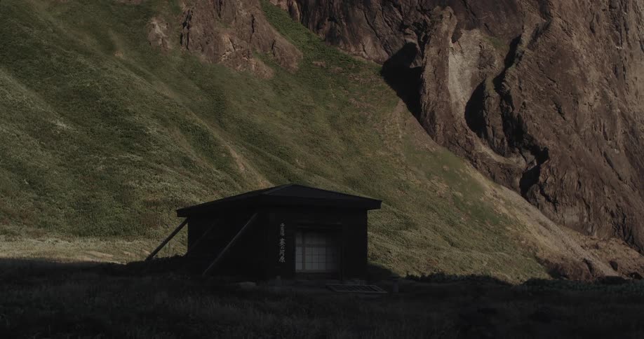 A small dark cabin sits nestled at the base of a large rugged grassy mountain slope under soft lighting