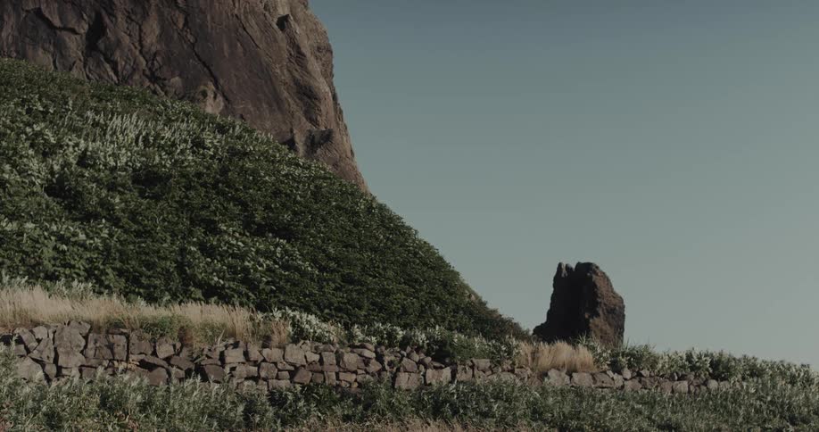 A static shot captures a massive cliff towering over a lush green hillside and a handbuilt stone wall with a jagged rock formation standing against a clear sky in a rugged coastal environment
