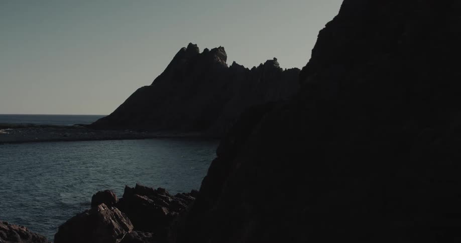 A wide shot reveals a rocky cove where dark crags meet deep blue water featuring a sharp mountain silhouette against a clear sky and a distant structure on the sparse shoreline