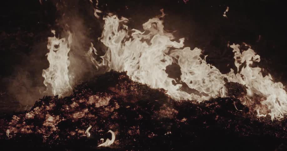 A large pile of dark smoldering material is engulfed in bright flickering flames against a dark background